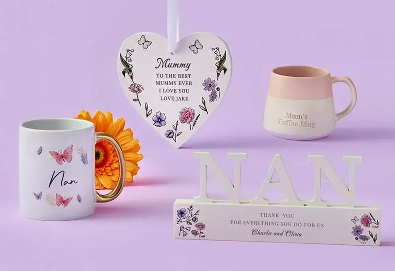 A pastel display of Nan and Mum-themed gifts arranged on a lavender background, including two decorative mugs, a heart-shaped hanging plaque with floral details, a NAN ornament, and an orange flower.